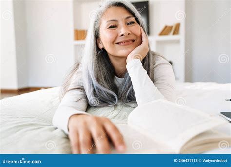 Grey Asian Woman Smiling And Reading Book While Lying On Bed Stock