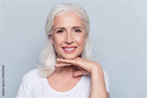 Charming Old Woman Holding Hand Under Chin With Beaming Smile Looking At Camera Over Gray