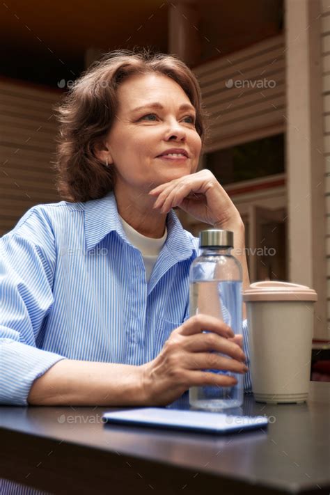 Adorable Mature Woman Sits At The Table In Outside Cafe With Paper