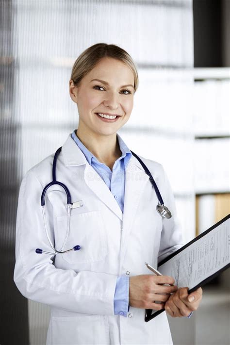 Friendly Female Doctor Standing And Holding Clipboard In Clinic