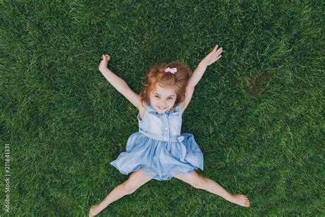 Smiling little cute child baby girl in light dress lie on green grass lawn, spreading hands and ...