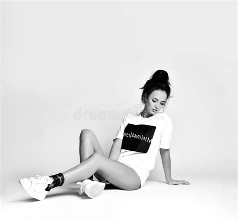 Beautiful Brunette Woman Sitting On Grey Background In Long White T Shirt And Sneakers Stock