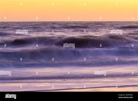 Two People Wearing Wetsuits And Carrying Boards Are Walking Along A Sandy Beach At Dusk As The