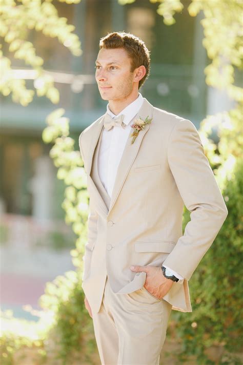 A Man Wearing A Tan Suit And Bow Tie Standing In Front Of Some Bushes