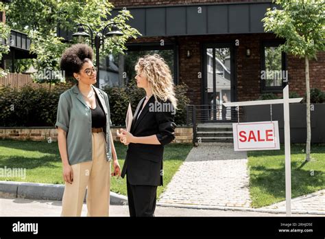 Blonde Real Estate Agent Talking To African American Client Near House And For Sale Signboard