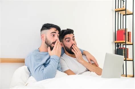 Pareja gay joven sorprendida que se sienta en la cama que mira el ordenador portátil Foto Gratis