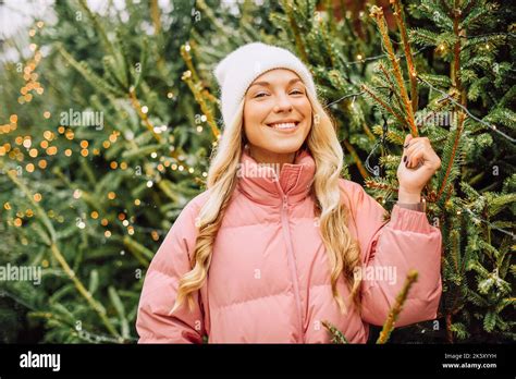A Cute Blonde Chooses A Christmas Tree For The New Year A Woman In A Hat And Pink Jacket Smiles