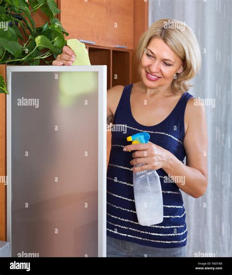 Mature Smiling Blonde Girl Cleaning Glass Door Of Furniture Stock Photo