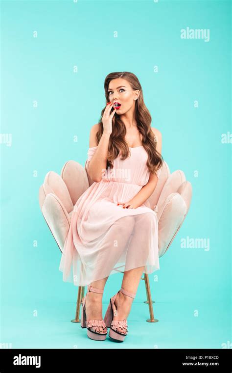 Full Length Photo Of Excited Woman 20s In Pink Dress Sitting In Soft Armchair With Legs Crossed