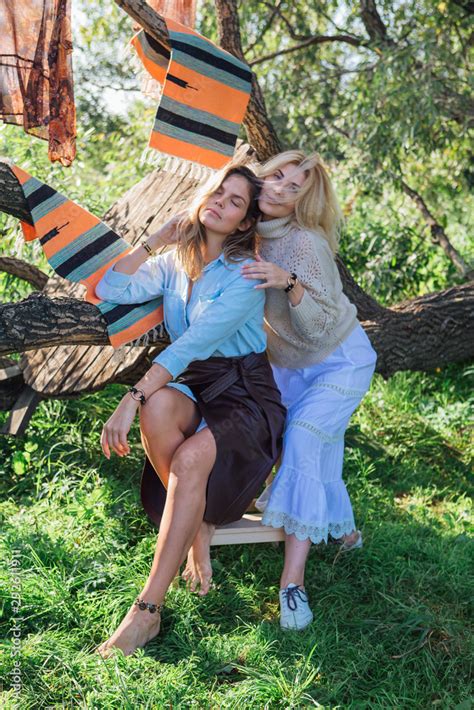 Portrait Of Two Beautiful Women Brunette And Blond Sitting On The Big Brunch Of Tree In A Field