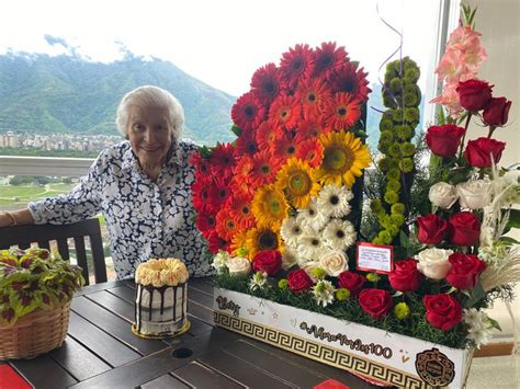 An Older Woman Sitting At A Table With Flowers And Cakes In Front Of