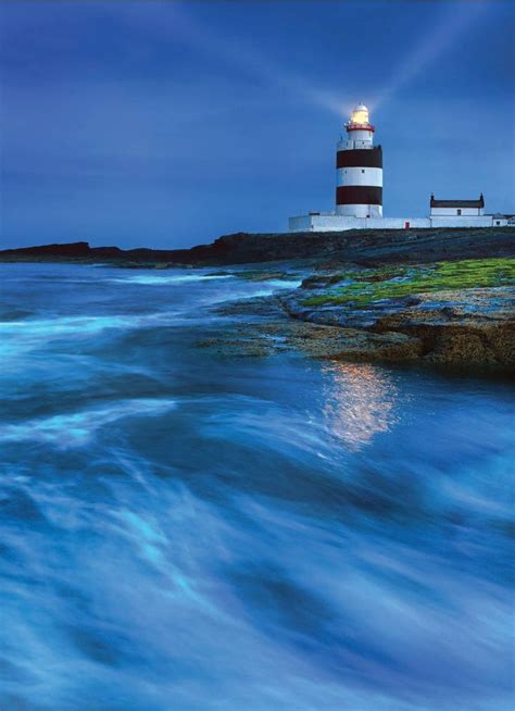 the light house is lit up at night by the water's edge with waves coming in