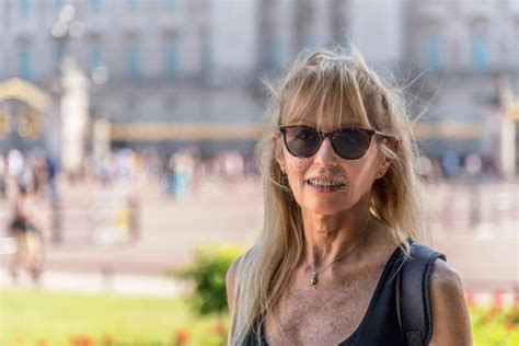Mature Blonde Woman With Glasses Looking At The Camera Behind Out Of Focus Buckingham Palace