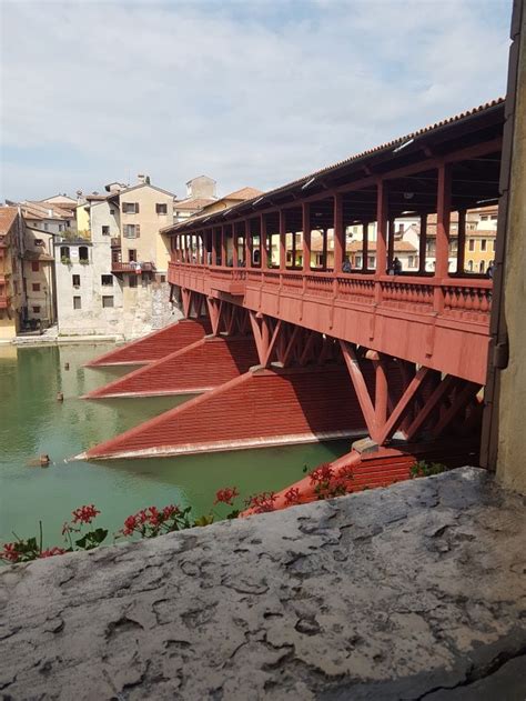 A Red Bridge Over Water With Buildings In The Background And Flowers