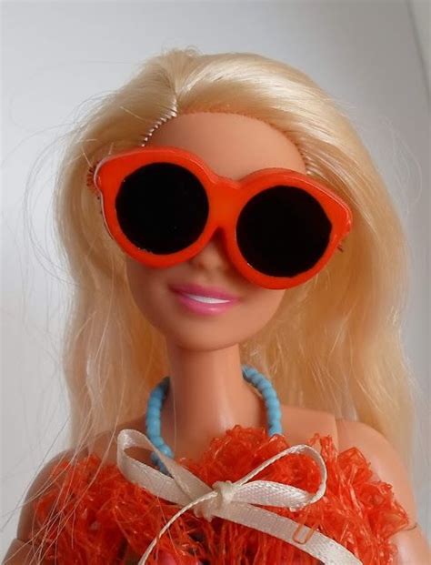 A Barbie Doll Wearing Bright Orange Sunglasses And A Blue Beaded Necklace With White Ribbons