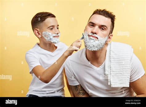 Father And His Son With Shaving Foam On Their Faces Are Shaving And