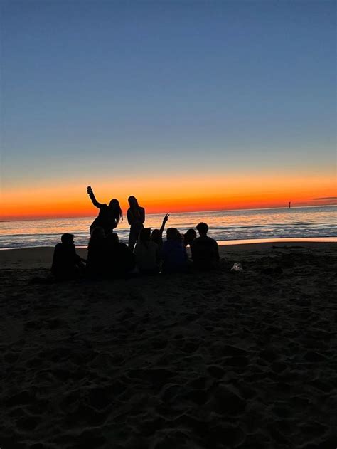 A Group Of People Sitting On Top Of A Beach Under A Colorful Sky At Sunset