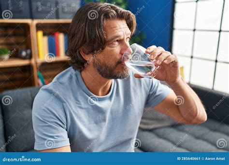 Middle Age Man Drinking Glass Of Water Sitting On Sofa At Home Stock