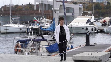 A Teenage Boy With Curly Hair Walks Along The Pier Along Moored Boats