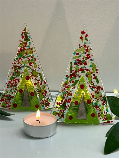 Two Small Christmas Trees Are Sitting Next To A Lit Candle On A White Surface With Green Leaves