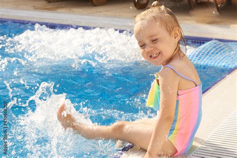 Little girl in rainbow swimsuit sit on the pool edge, swinging her feet ...