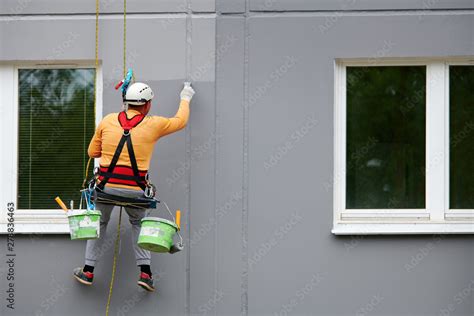 Worker Hanging On Rope And Paints Building Wall With Roller Painter