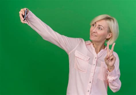 Souriante jeune femme slave blonde faisant signe de paix et prenant selfie isolé sur fond vert