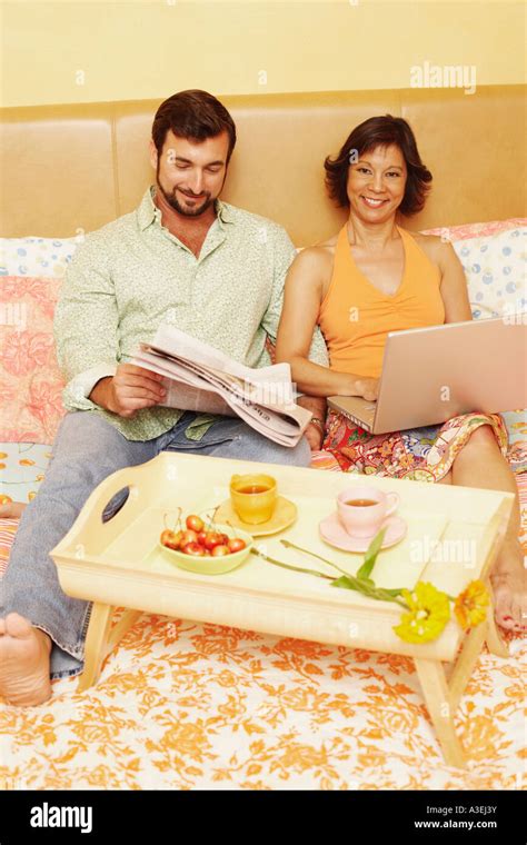 Mature Man Reading A Newspaper With A Mature Woman Using A Laptop On The Bed Stock Photo Alamy