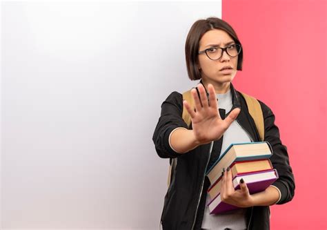 안경을 착용하고 흰색 벽 앞에 서있는 책을 들고 불쾌한 젊은 학생 소녀 복사 공간이 분홍색 배경에 고립 된 몸짓 무료 사진