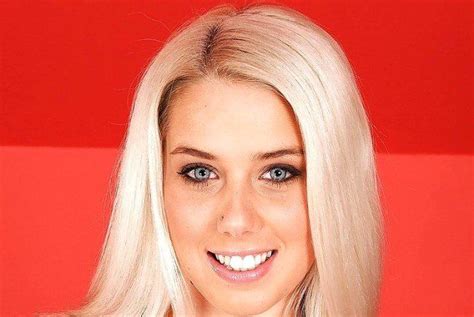 A Woman With Blonde Hair And Blue Eyes Smiles At The Camera While Standing In Front Of A Red