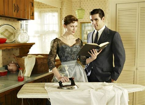 A Man And Woman Standing In A Kitchen Next To An Ironing Board With A