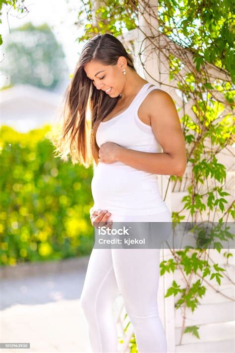 Smiling Pregnant Woman Standing In Garden Holding Hands On Belly Stock