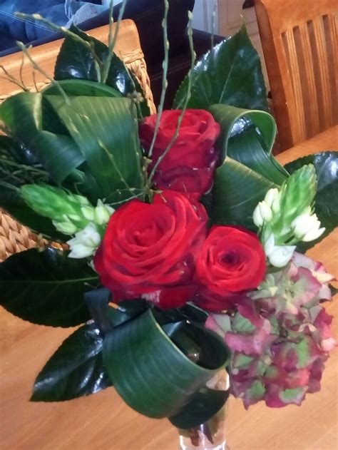 A Vase Filled With Red Roses And Greenery On Top Of A Wooden Dining