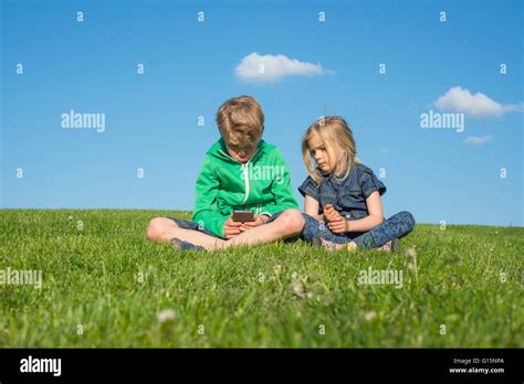 Glückliche blonde Kinder mit Smartphone Film oder Spiel sitzen auf dem Rasen Bruder und