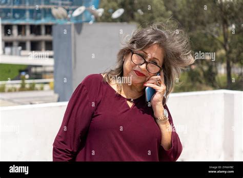 Senior Latina Woman With Glasses Talking On Cell Phone On A Terrace With Trees And Buildings