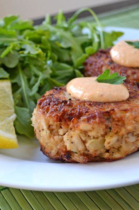 Two Crab Cakes On A Plate With Greens And Lemon Wedges