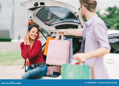 Caucasian Man Gives Shopping Bags To Asian Woman Sitting On Car