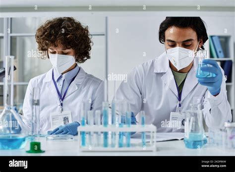 Two Young Clinicians In Labcoats Respirators And Gloves Working With