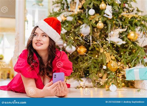 A Woman In Santa Claus Cap Under The Christmas Tree With A Smartphone