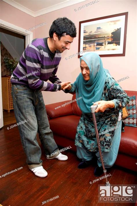 South Asian Son Helping His Mother Get Up From Sofa Stock Photo