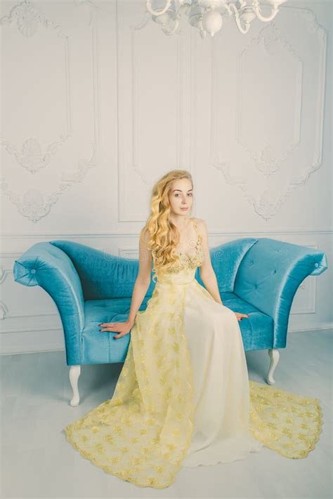 Retrato Integral De Un Blonde Joven Hermoso En Un Vestido Largo Del Oro En El Sof Estudio Alta