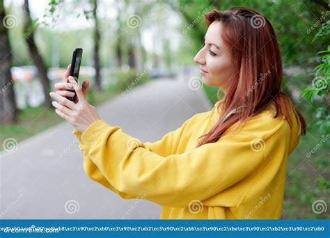 A Beautiful Girl In A Yellow Hoodie Stands Sideways Holding A Phone Taking A Selfie Or Looking