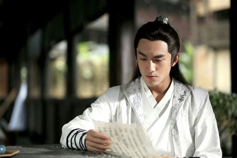 A Man Sitting At A Table Reading A Piece Of Paper While Wearing A White