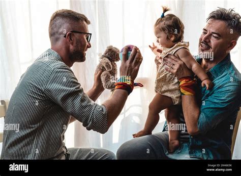 Male Gay Couple With Adopted Baby Girl At Home Two Handsome Fathers Playing With Their