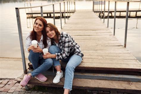 Cute Lesbian Couple Sits Together Near The Lake With Cups Of Drink In Hands And Embracing Each