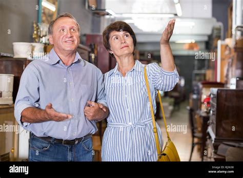 Mature Man With His Wife Are Choosing Furniture In The Antique Shop Focus On Both Persons Stock
