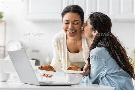 Happy Lesbian Woman Kissing Cheek Of Cheerful Multiracial Girlfriend With Croissants Stock Photo