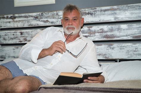 Mature Gray Haired Man With Beard And Eyeglasses In Hand In Pajamas Lying In Bed Looking At