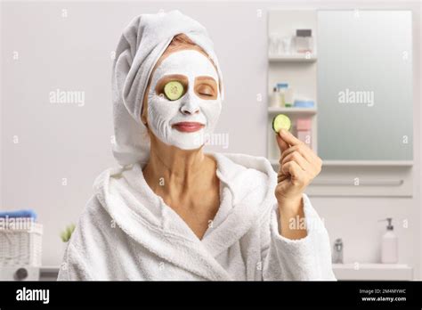 Mature Woman With A Towel On Her Head And A Face Mask Putting Cucumber On Eyes Inside A Bathroom