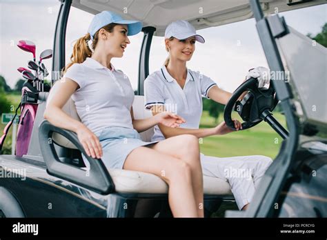 Smiling Female Golfers In Caps Riding Golf Cart At Golf Course Stock